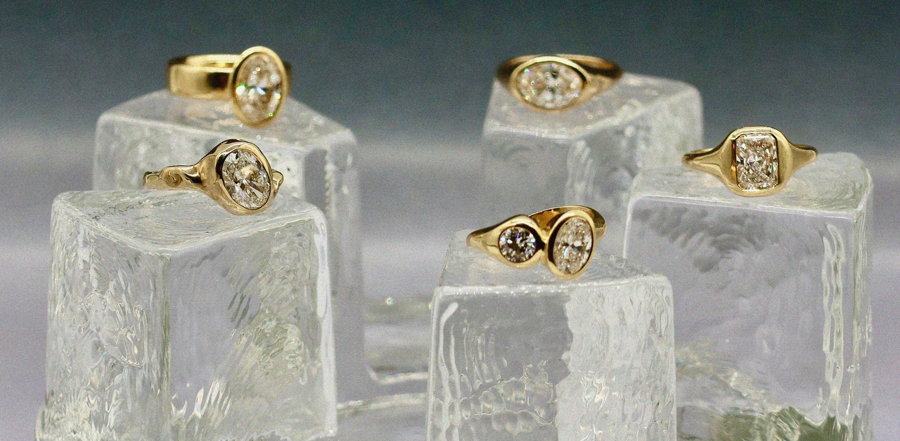 Five gold rings with round-cut clear gemstones displayed on textured glass blocks against a dark gray background.