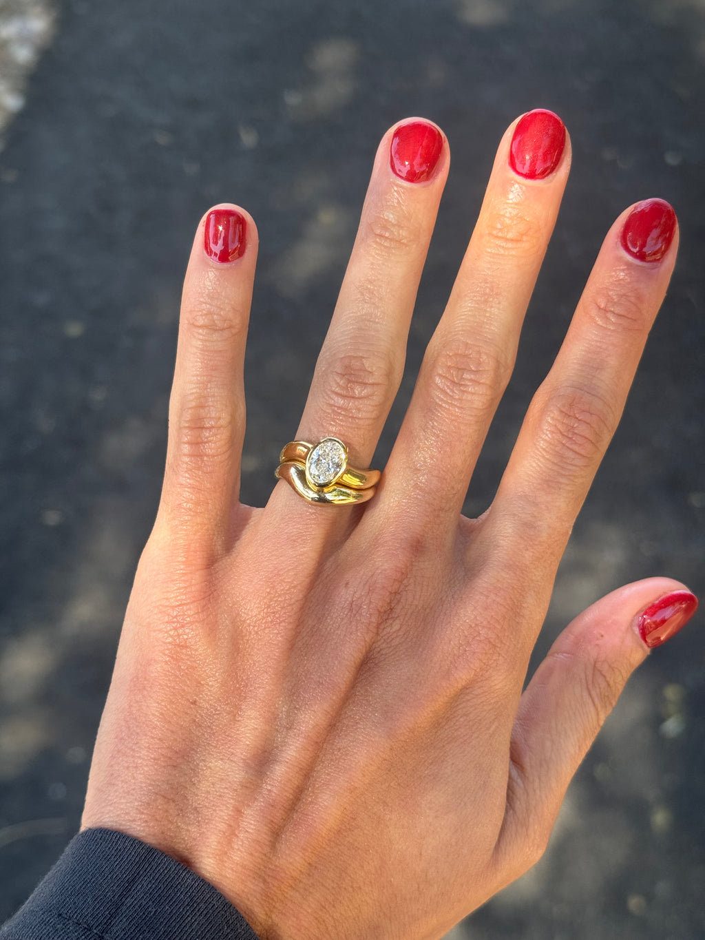 Gold ring asymmetric design with oval diamond and matching paired wedding band, on the finger, blurred outdoor background.