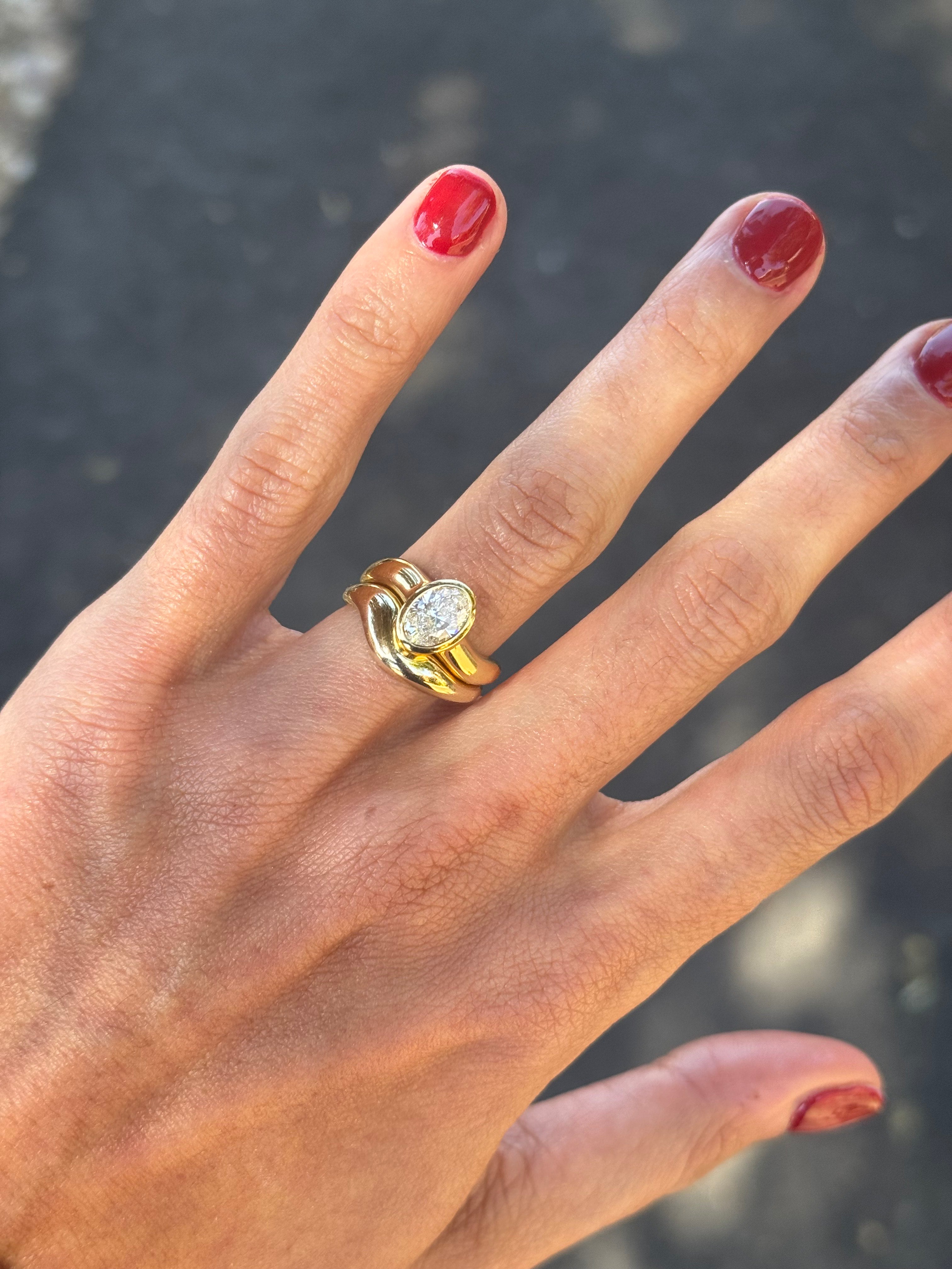 Gold ring asymmetric design with oval diamond and matching paired wedding band, on the finger, blurred outdoor background.