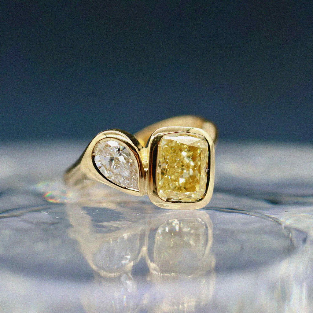 A yellow gold ring featuring a cushion-cut yellow diamond and a smaller marquise-cut clear diamond, displayed on a reflective clear surface against a dark blue background.