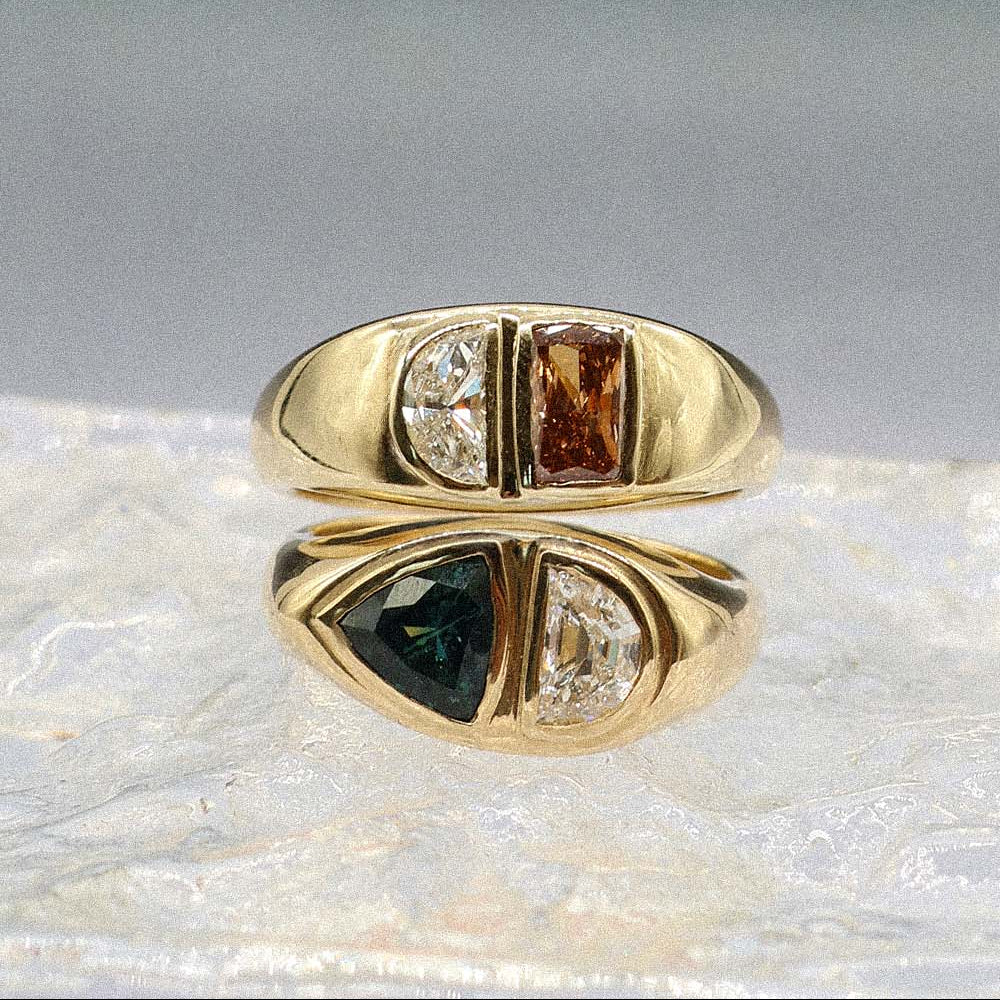A gold ring with three marquise-cut gemstones set in a row, displayed on a textured white surface with its reflection visible. The center stone is a warm brown, and the outer stones are one dark green and one clear.