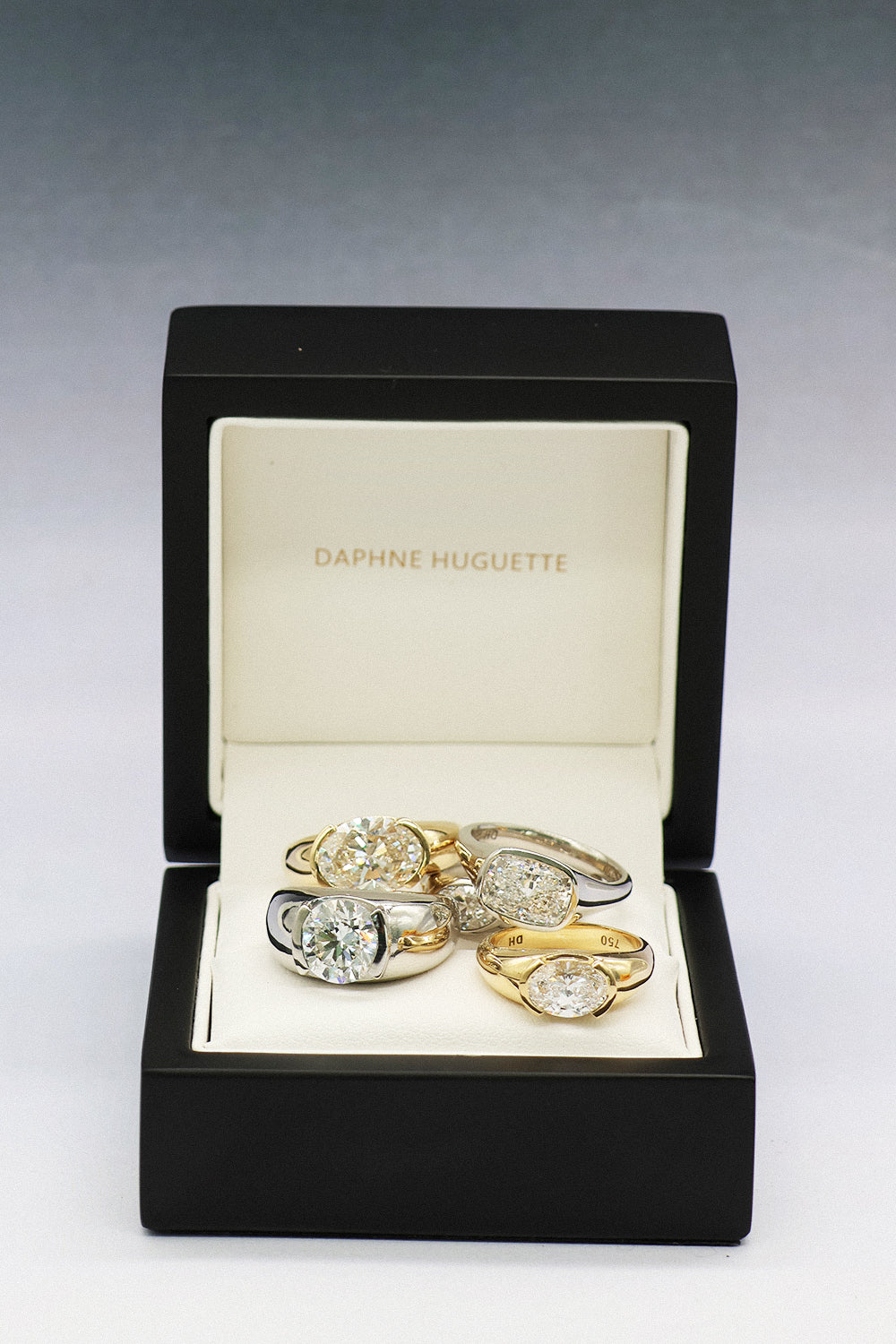 A set of five Daphne Huguette rings displayed inside an open black box with a cream interior. The rings feature yellow gold and white gold metals with clear diamonds.