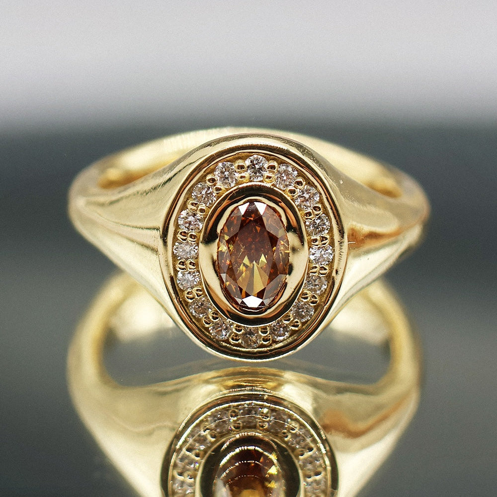 A gold signet-style ring with an oval faceted brown center stone surrounded by a halo of small clear stones, shown on a reflective dark surface against a soft gray background.