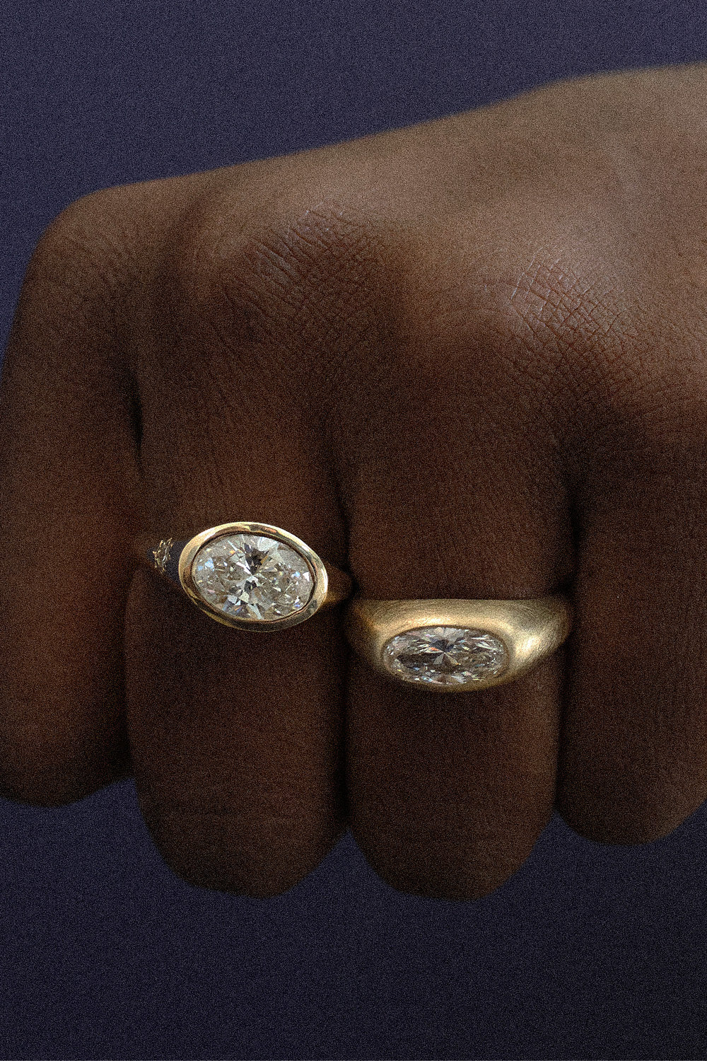 Two gold signet rings with oval-cut clear gemstones worn on a person's fingers, set against a dark navy background.