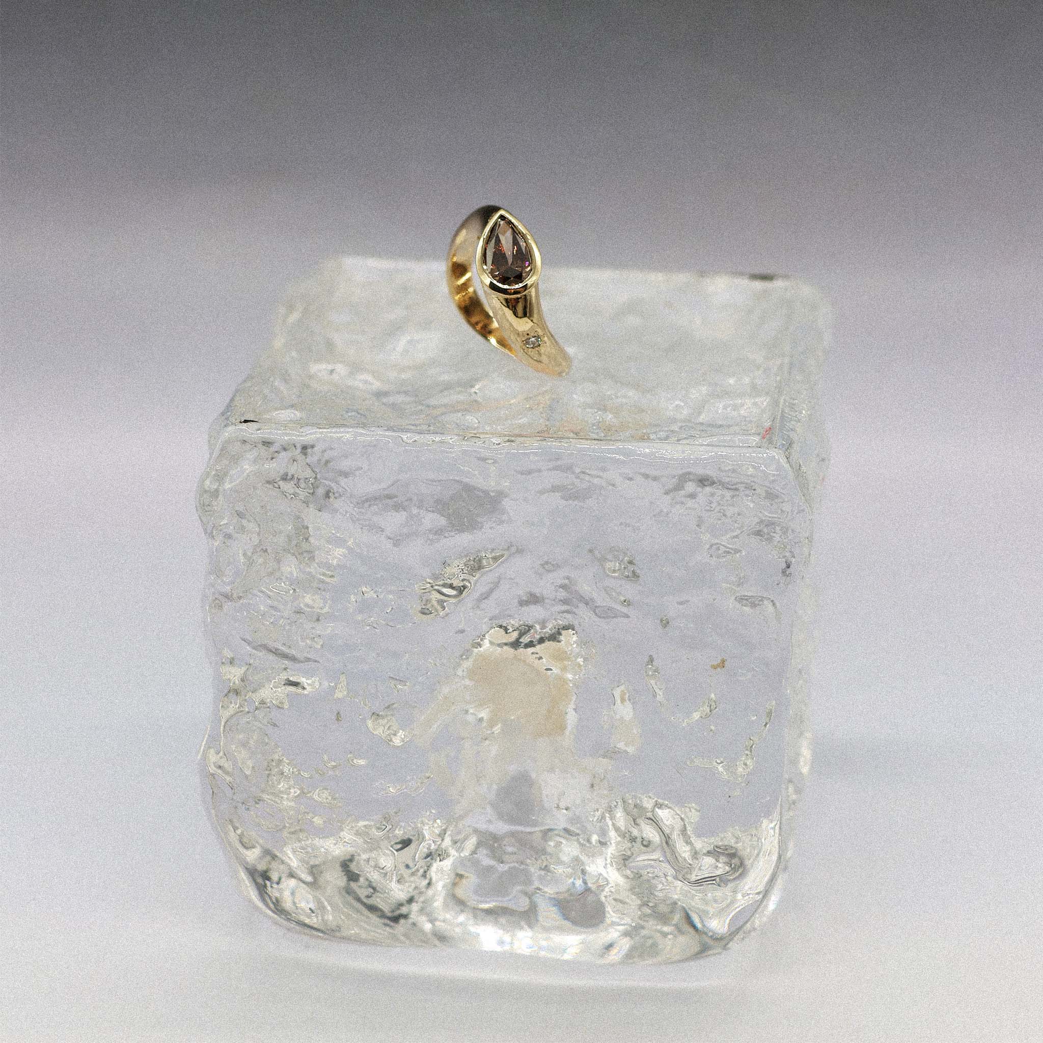 A gold ring with a pear-cut brown diamond displayed on top of a clear, textured glass block against a neutral gray background.