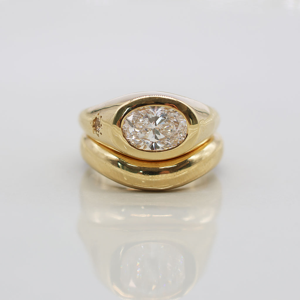 A yellow gold ring set with a round brilliant-cut diamond in an oval bezel, featuring a smaller round diamond on the side, displayed on a reflective white surface.