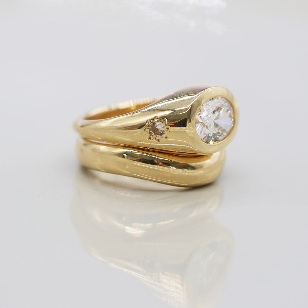 A yellow gold ring set featuring a large round brilliant-cut diamond on the main band and a smaller round diamond on a side post, photographed on a reflective white surface.