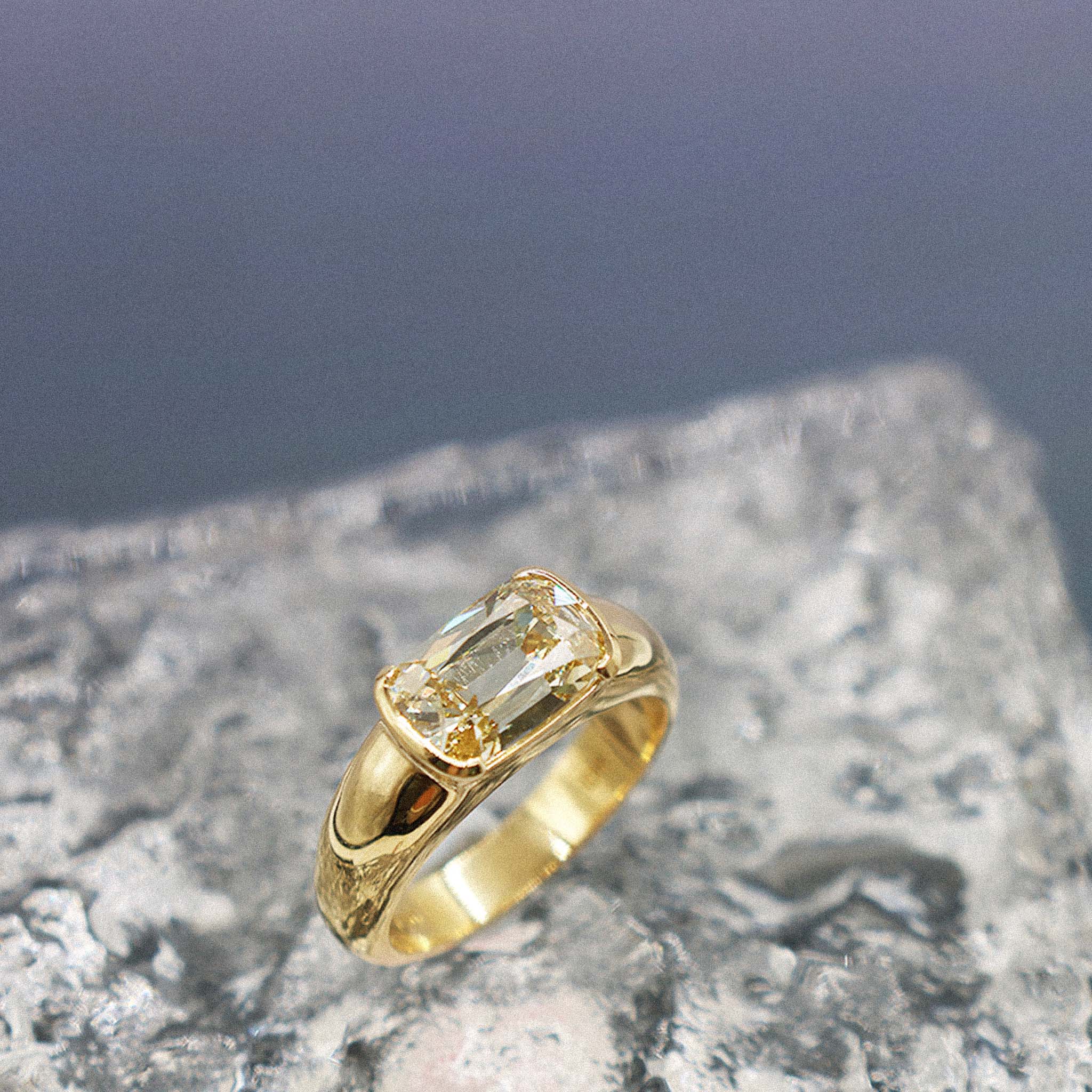 A yellow gold ring featuring an antique-cut yellow diamond, displayed on a textured gray stone surface with a soft-focus blue-gray background.
