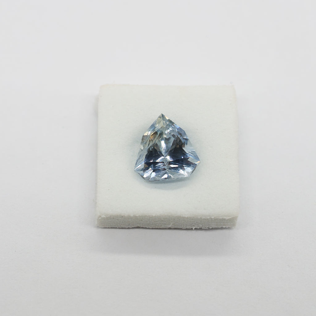 A light blue triangular faceted sapphire displayed on a small white foam block against a plain light background.