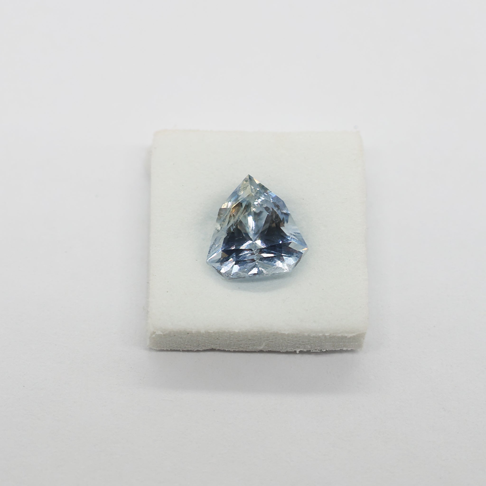 A light blue triangular faceted sapphire displayed on a small white foam block against a plain light background.