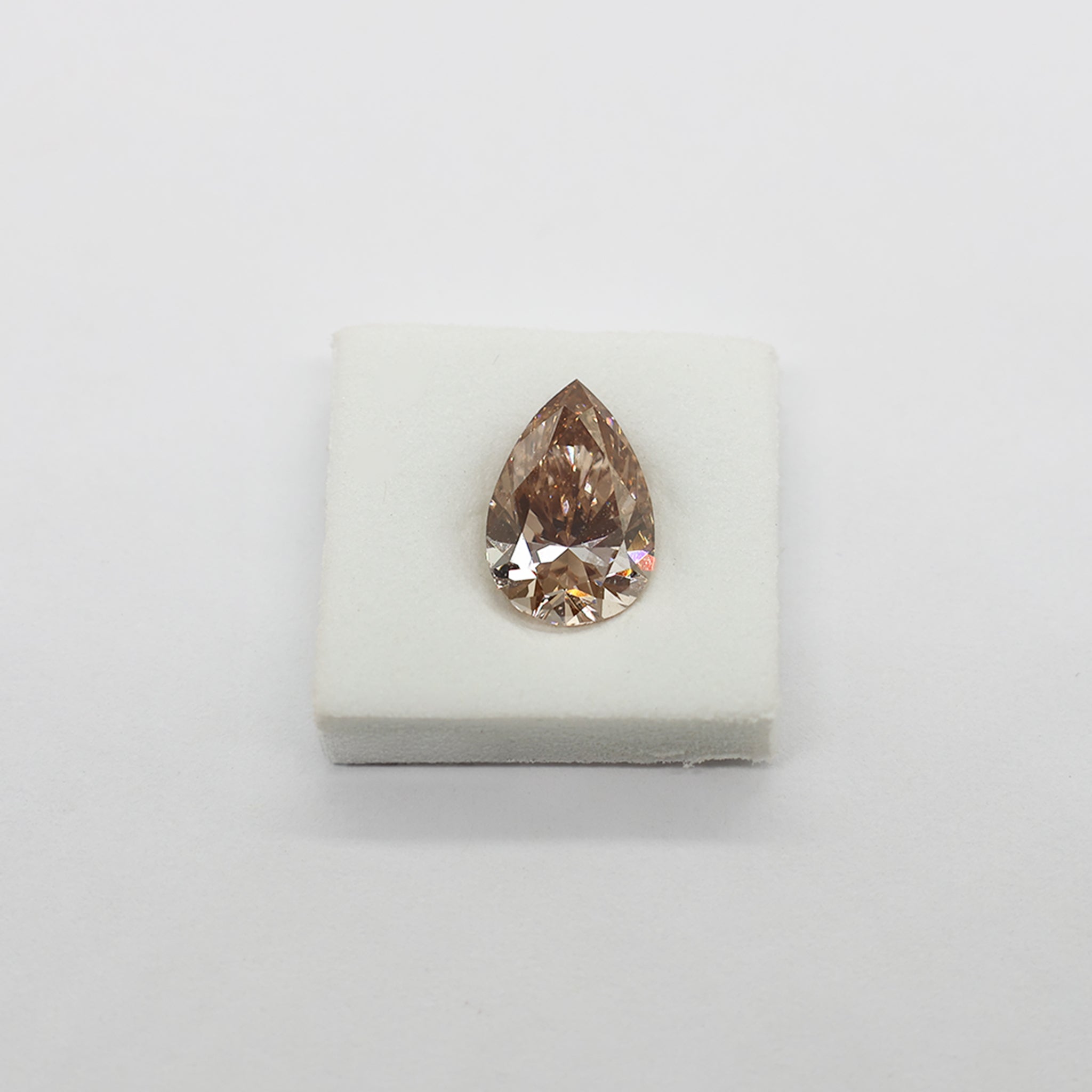 A single pear-cut peach-colored diamond displayed on a small white square cushion against a plain light background.