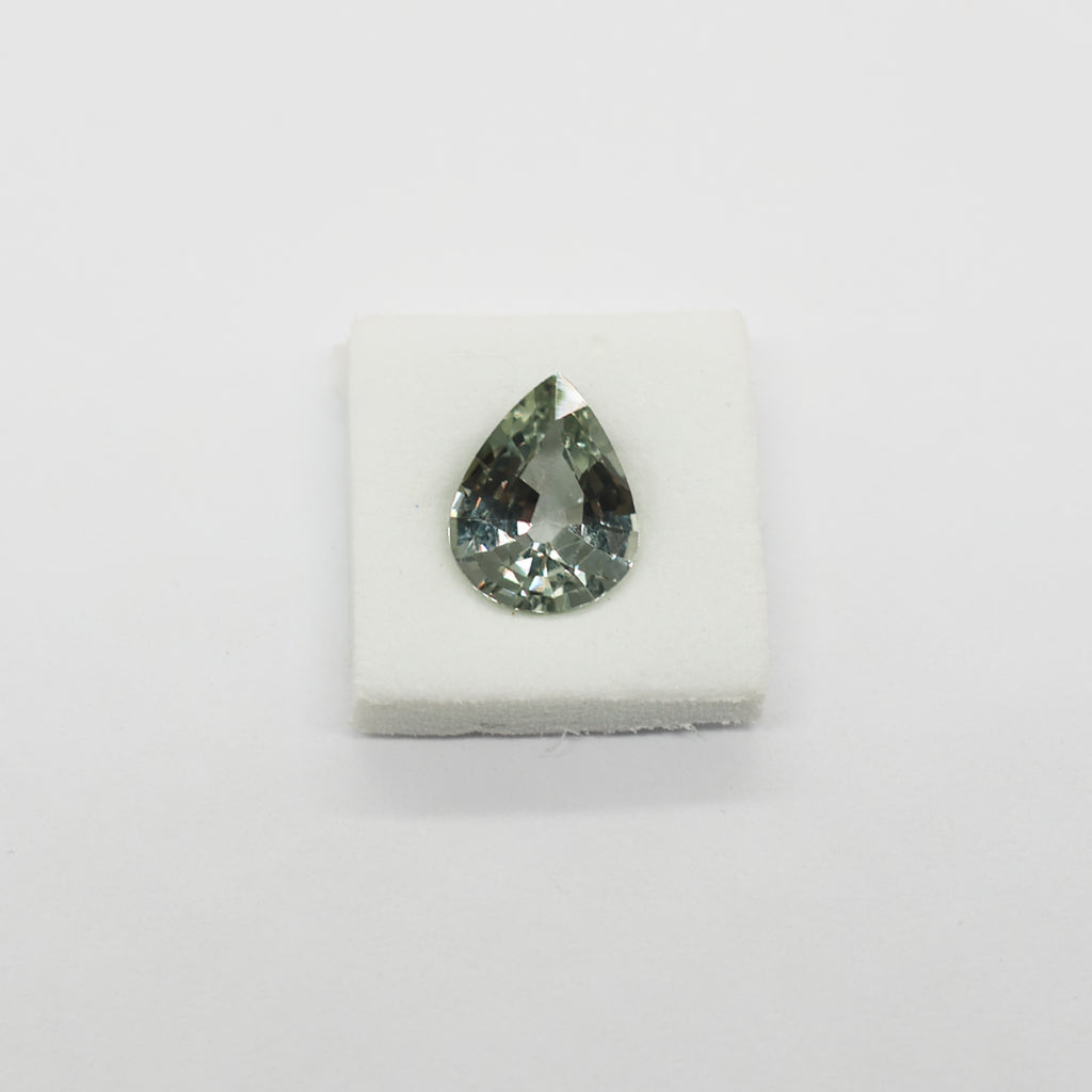 A teardrop-shaped faceted sapphire with a greenish-gray hue, displayed on a small white foam block against a plain white background.