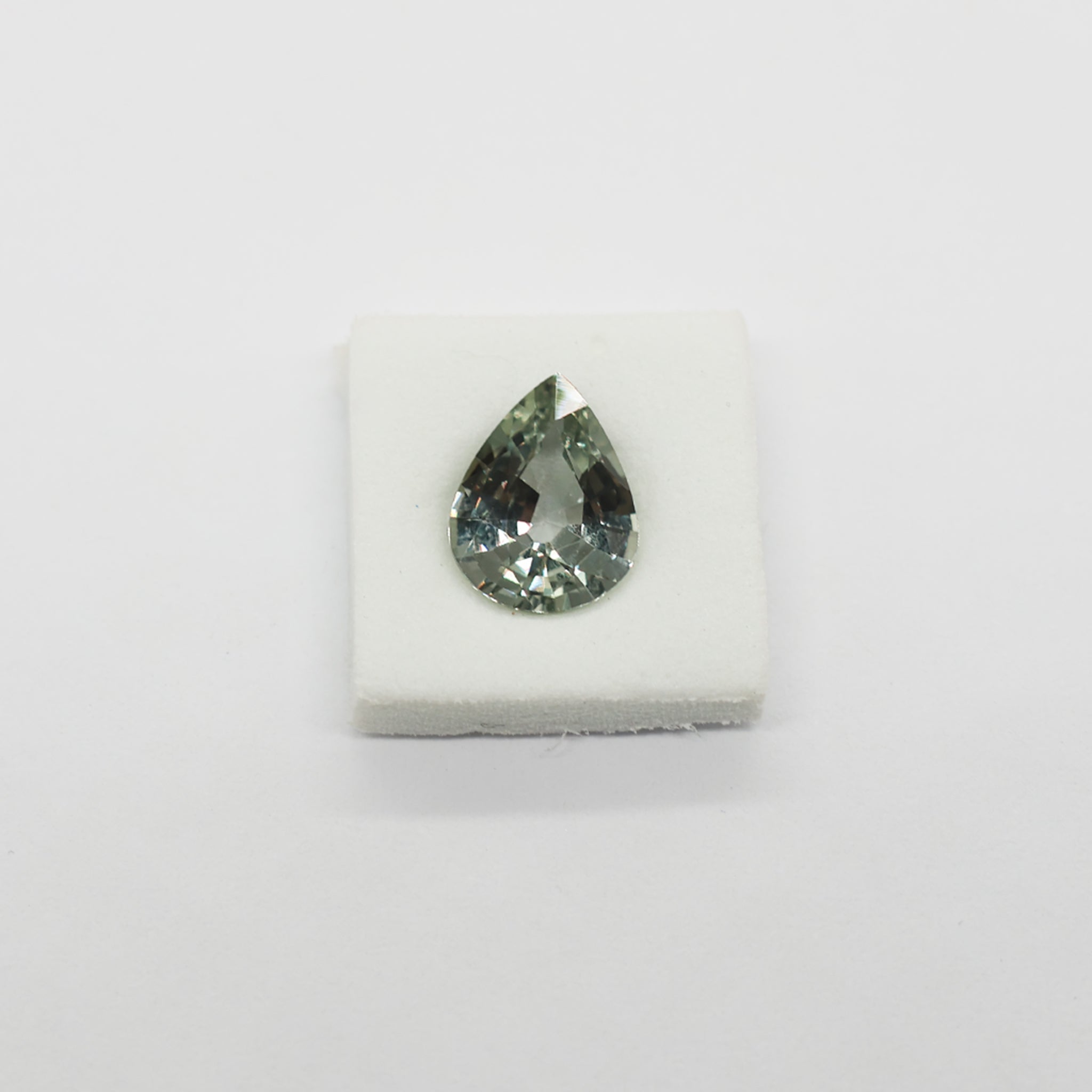 A teardrop-shaped faceted sapphire with a greenish-gray hue, displayed on a small white foam block against a plain white background.